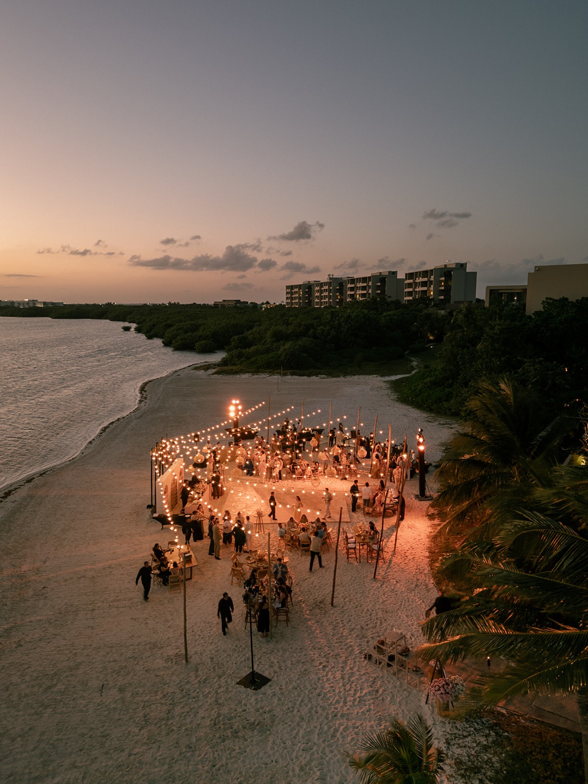 Aerial view of a bespoke luxury beach wedding setup at sunset with circular seating and ambient lighting.